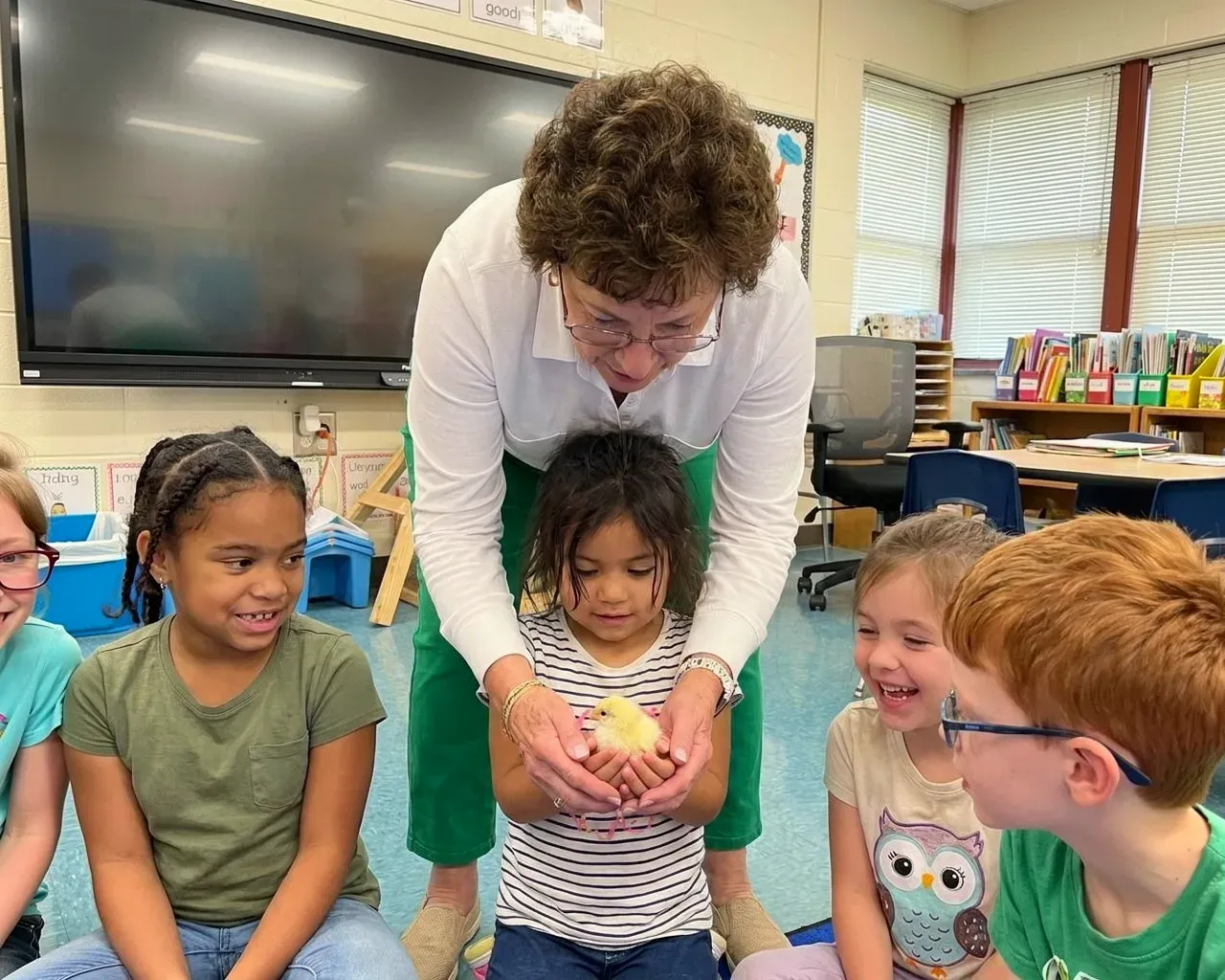 A teacher helping a young student hold a chick while classmates watch